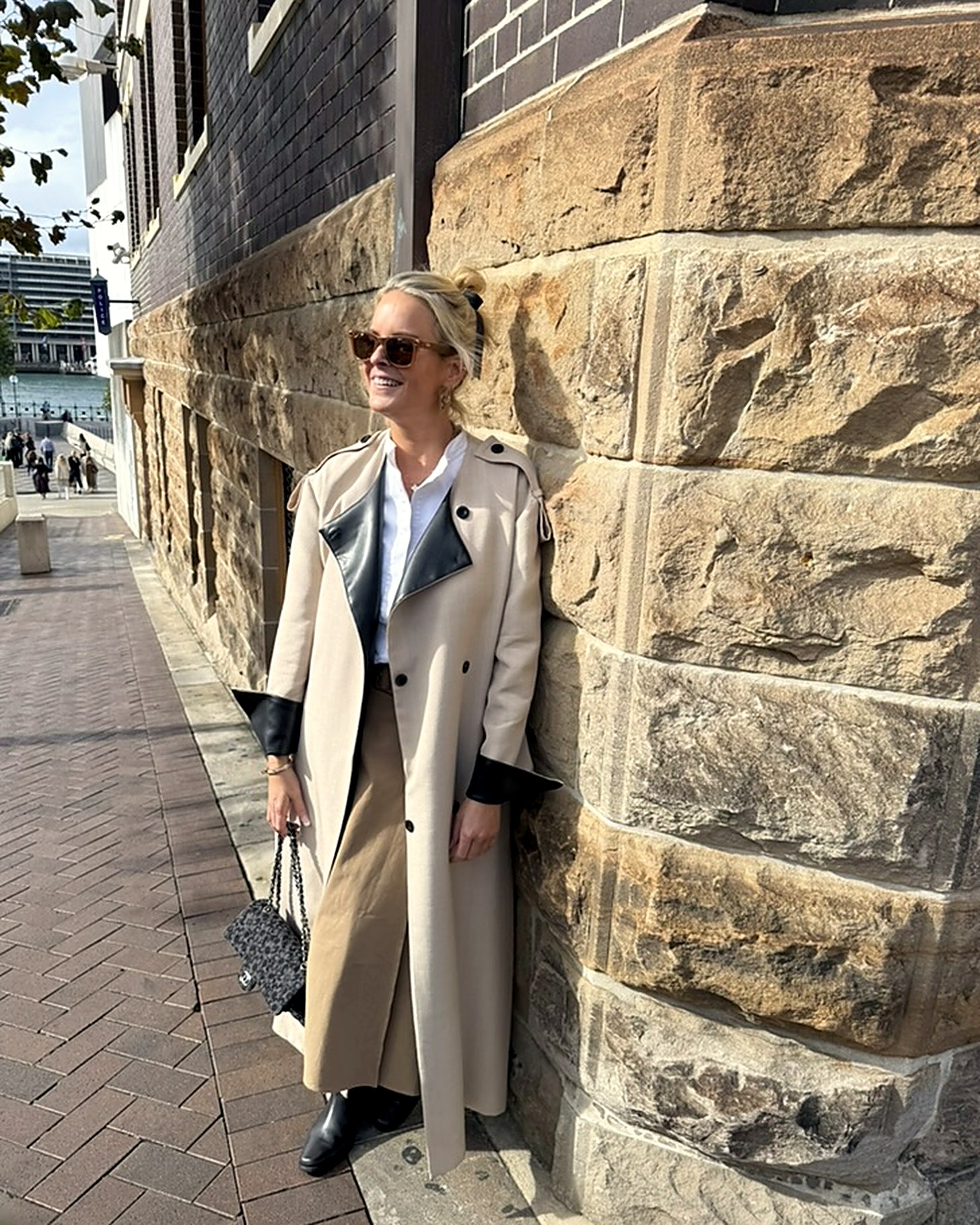 The artist leaning against a sandstone wall in Sydney, wearing a cream trench coat